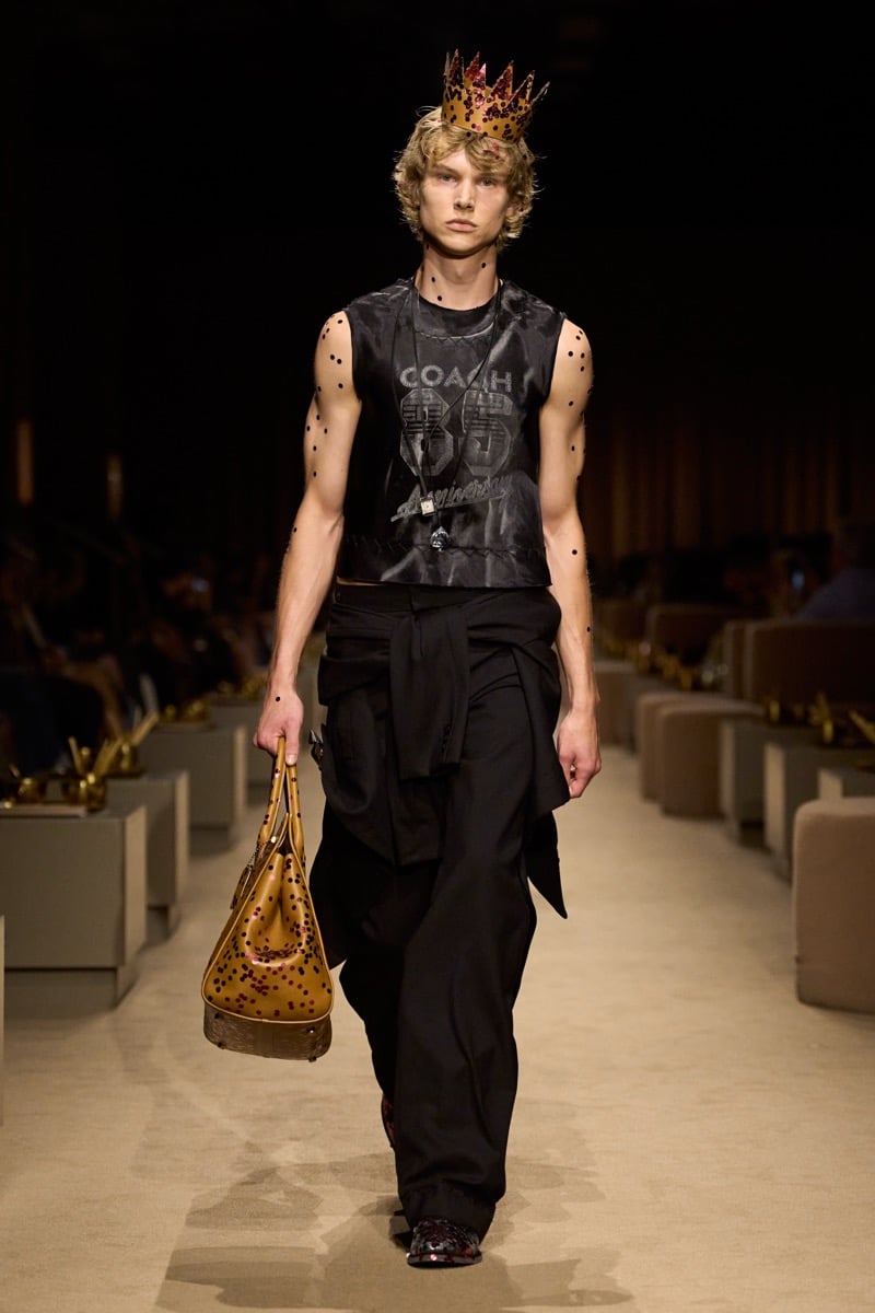 A model in a paper crown and a black Coach 85th-anniversary graphic tank with wide black trousers, a dot-print tan bag in one hand, for Coach's spring 2027 collection