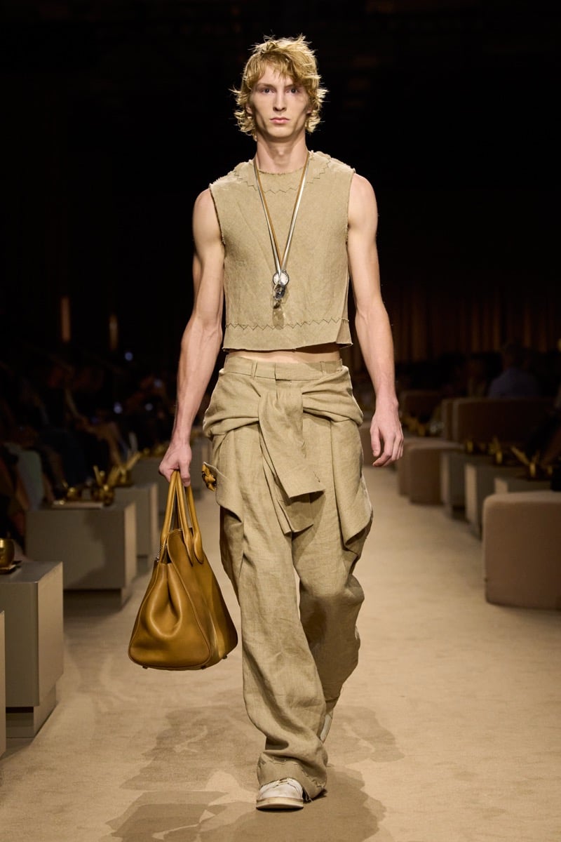 A model in a cropped beige knit tank and slouchy khaki linen trousers with fabric tied at the waist, a tan leather bag in one hand, for Coach's spring 2027 collection