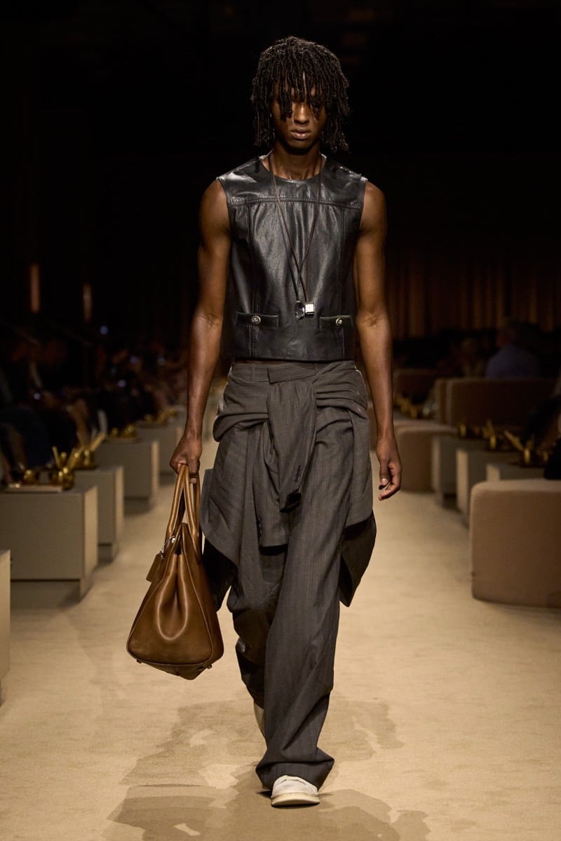 A model in a cropped black leather vest and slouchy pinstripe trousers with a shirt knotted at the waist, a brown leather tote in one hand, for Coach's spring 2027 collection