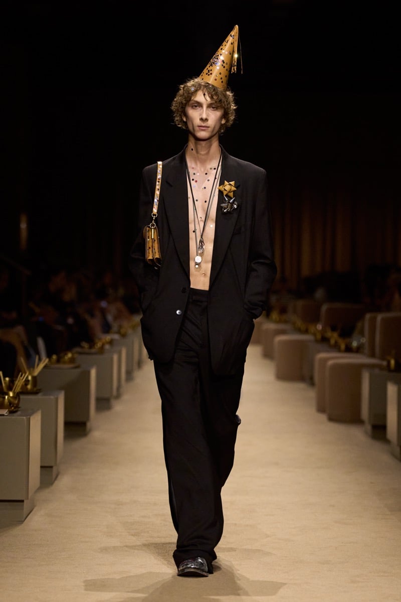 A model in a gold cone party hat and an oversized black suit worn open over a bare chest, a tan shoulder bag at his side, for Coach's spring 2027 collection