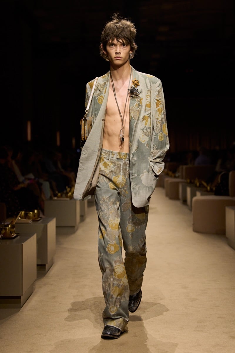 A model in a floral-jacquard suit worn open over a bare chest, with pendant necklaces and a small shoulder bag, for Coach's spring 2027 collection