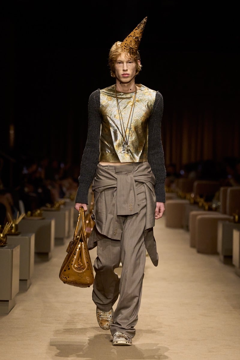 A model in a gold cone party hat and a gold lame top with contrast knit sleeves, slouchy taupe pinstripe trousers and a jacket tied at the waist, an embellished tan bag in hand, for Coach's spring 2027 collection
