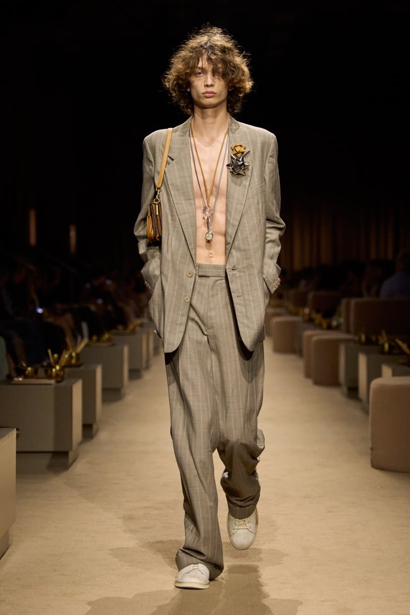 A model in an oversized gray pinstripe suit worn open over a bare chest, with layered pendant necklaces and a tan leather shoulder bag, for Coach's spring 2027 collection
