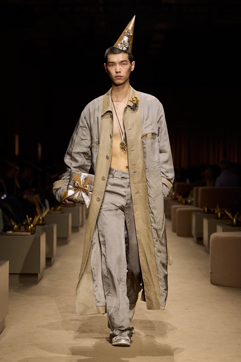 A model in a gold cone party hat and a gray silk trench coat over matching trousers, holding a wrapped gift, for Coach's spring 2027 collection