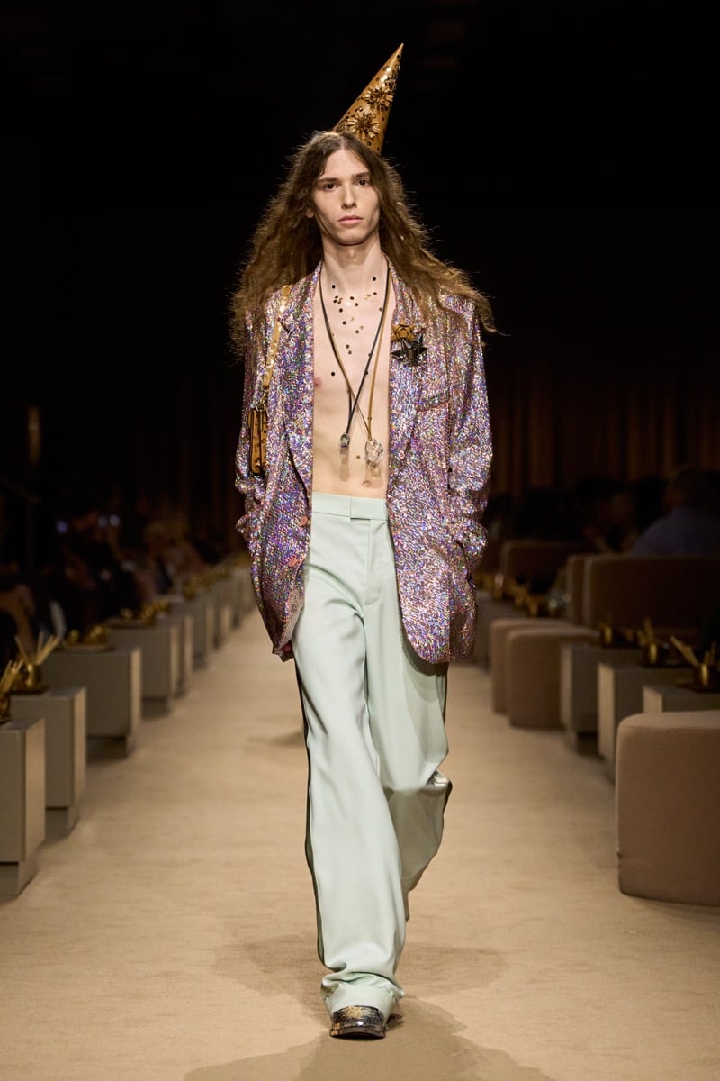 A model with long hair in a gold cone party hat and an iridescent sequined blazer worn open over a bare chest with pale green wide-leg trousers, for Coach's spring 2027 collection