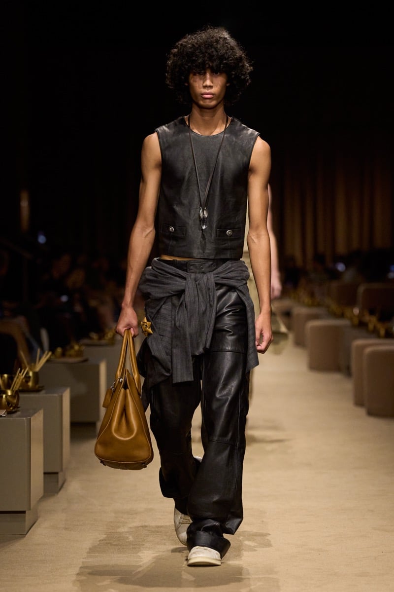 A model in a cropped black leather vest over a dark tee with black leather trousers and a shirt tied at the waist, a tan leather bag in hand, for Coach's spring 2027 collection