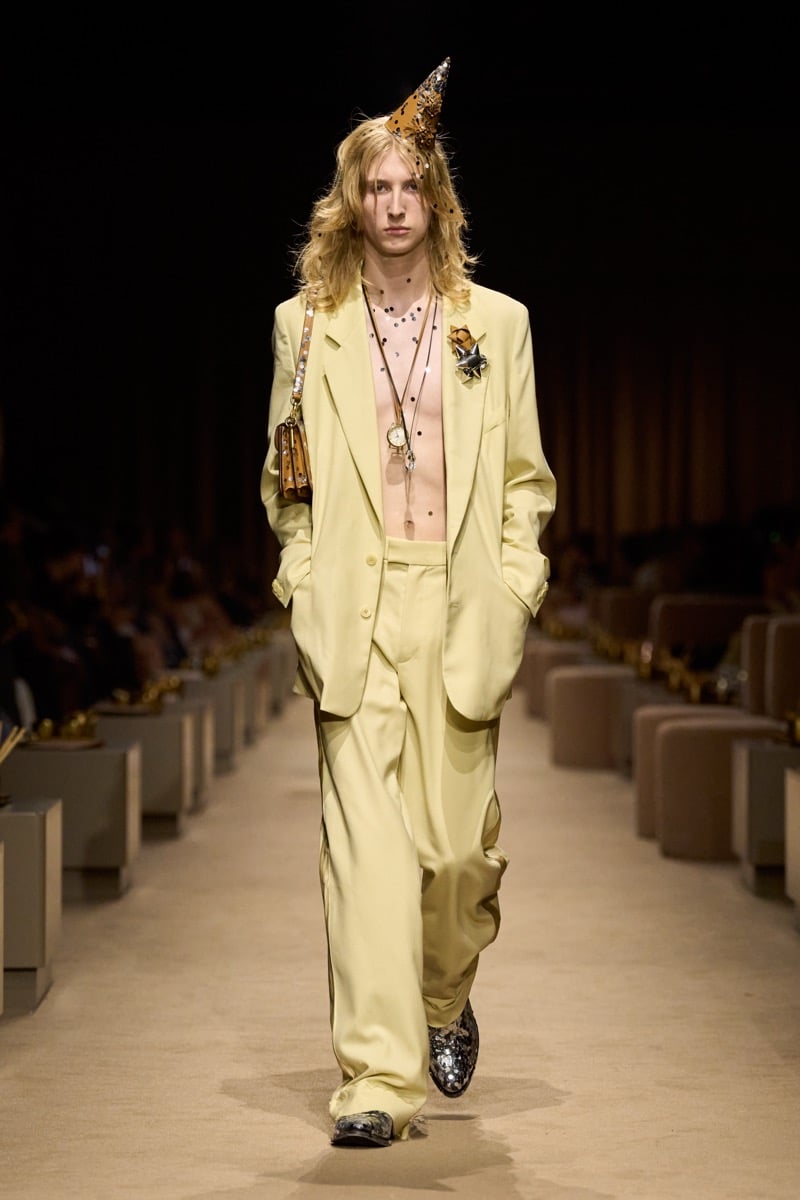 A model in a gold cone party hat and an oversized pale yellow suit worn open over a bare chest, with a small shoulder bag, for Coach's spring 2027 collection
