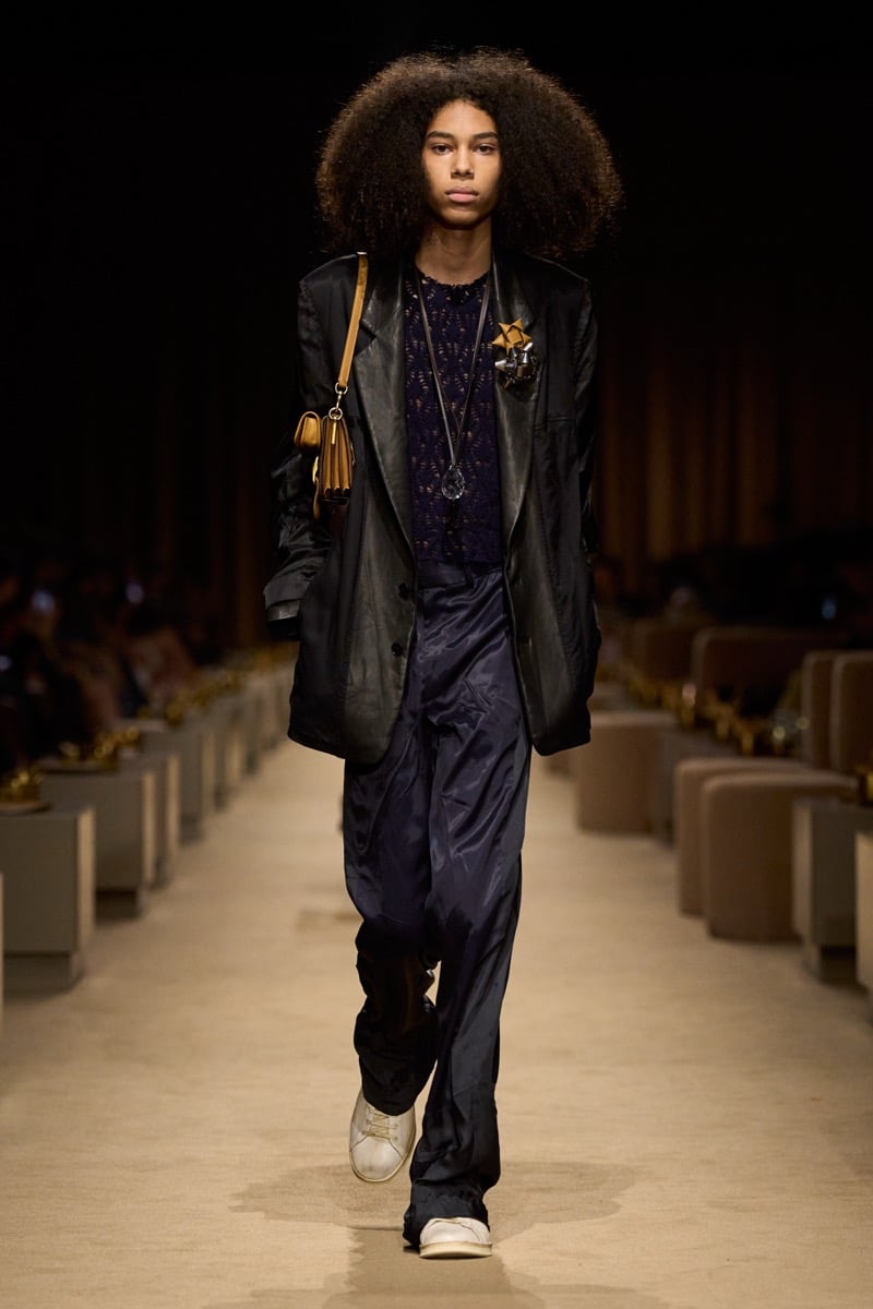 A model in a sheer dark blazer over a lace knit top with slouchy satin trousers and a small tan shoulder bag, for Coach's spring 2027 collection