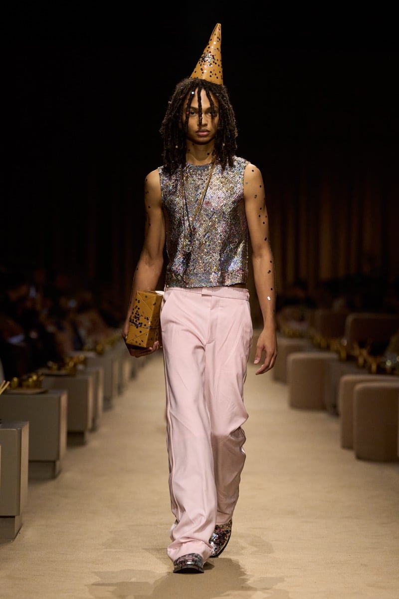 A model in a gold cone party hat and a silver sequined tank with pale pink wide-leg trousers, holding a small wrapped gift, for Coach's spring 2027 collection
