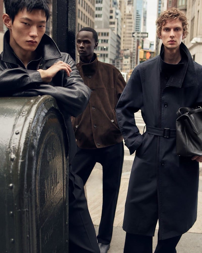 Hoyong Kim in a leather jacket stands beside Malick Bodian in a brown suede jacket and Leon Dame in a belted trench with a leather bag on a New York street, for COS's fall 2026 campaign