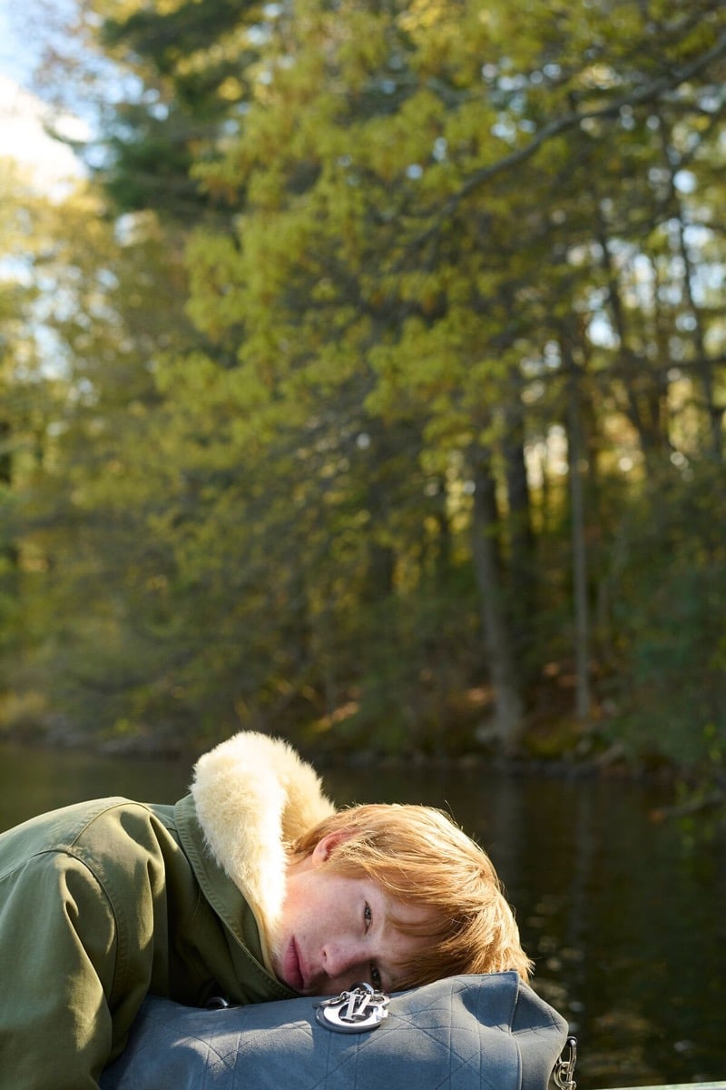 Cullan Sturdevant in a green parka with a shearling collar rests against a quilted Dior bag by a wooded lake