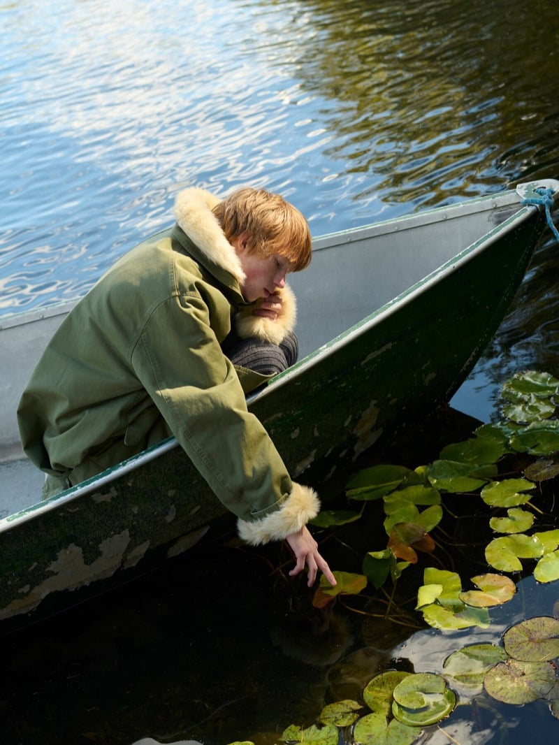 Cullan Sturdevant in a green parka with shearling cuffs leans over the side of a rowboat to touch lily pads