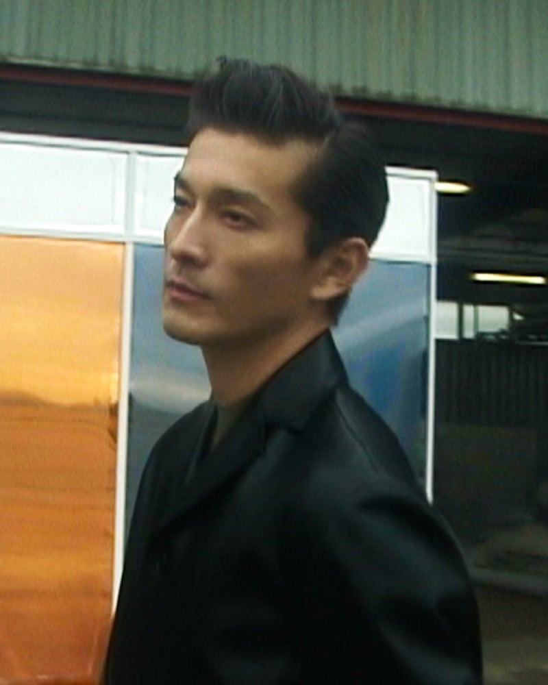 Daisuke Ueda seen in profile with slicked-back hair, wearing a boxy black leather jacket in front of orange and blue foil-wrapped panels outside an airport hangar, for the Jil Sander fall 2026 campaign