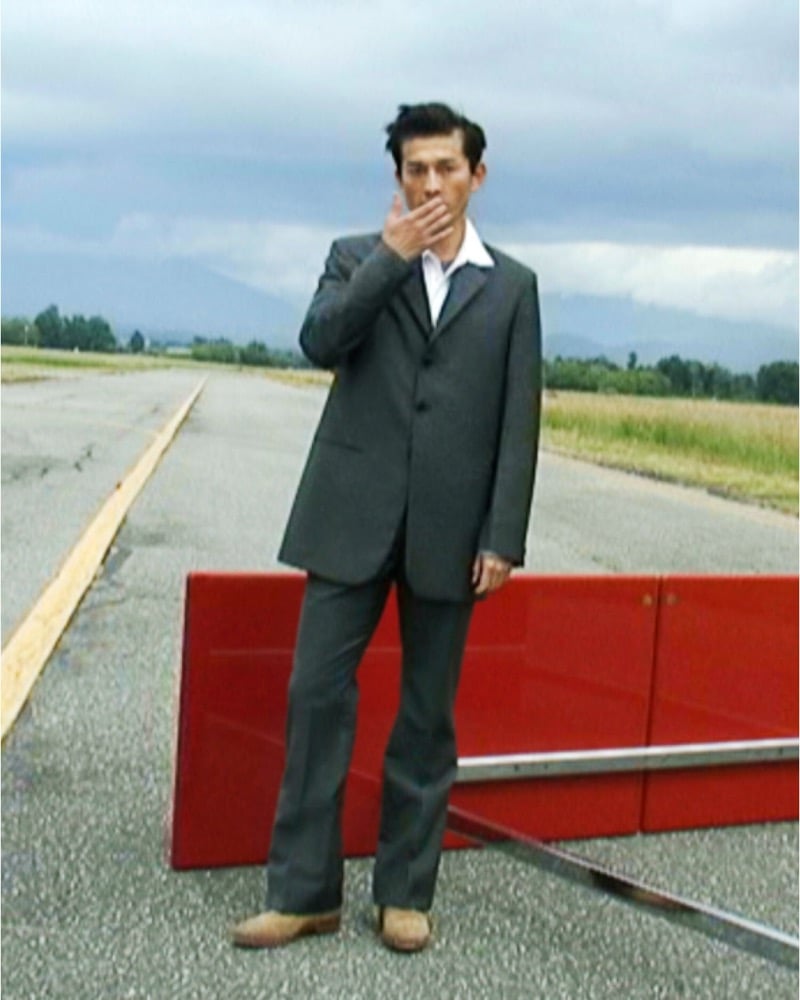 Daisuke Ueda stands on a cracked runway in a loose gray single-breasted suit with a white shirt and tan suede boots, a red lacquered panel and a metal pole on the ground behind him, for the Jil Sander fall 2026 campaign