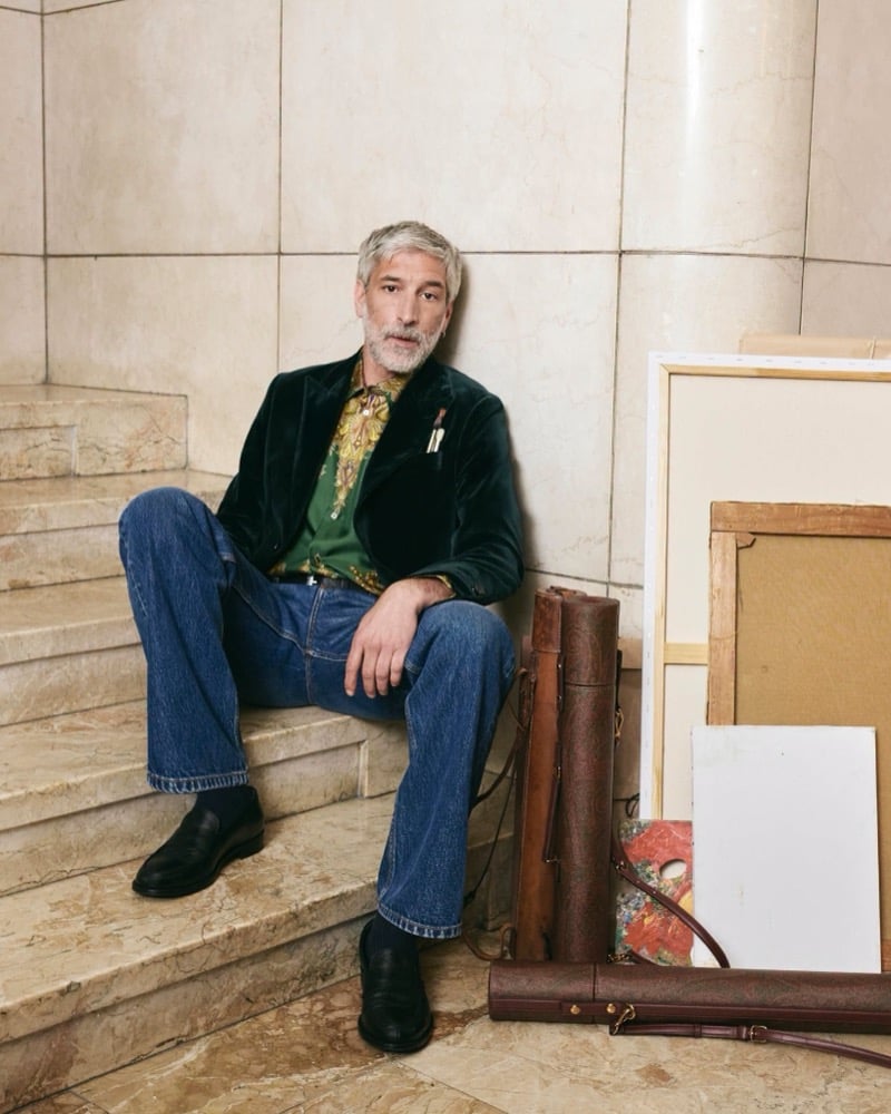 Duccio Maria Gambi sits on marble steps in a dark velvet blazer over a gold-toned paisley-print shirt and green knit, with blue jeans and black loafers, leather artist portfolio tubes and blank canvases propped beside him, for the Etro fall 2026 campaign