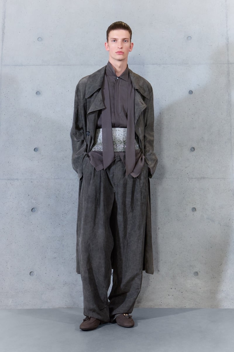 A model in a long coat over a pleated shirt, narrow scarf, and embellished sash, with wide-leg trousers, for Emporio Armani's spring-summer 2027 collection