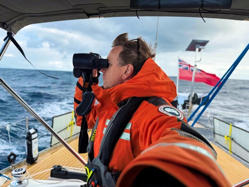 Ewan McGregor looks through binoculars aboard a sailboat in orange Canada Goose sailing gear with reflective strips and a harness