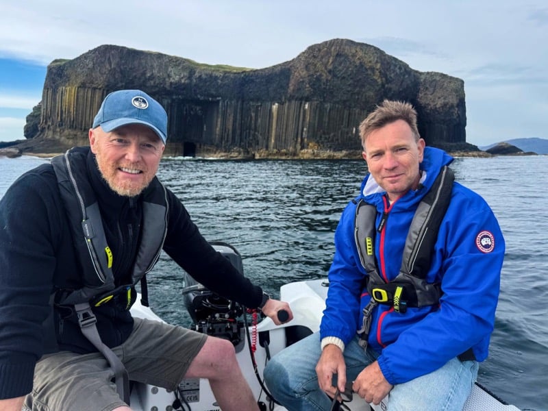 Ewan McGregor in a blue Canada Goose jacket and life vest sits in a dinghy beside a friend near the basalt cliffs of Staffa