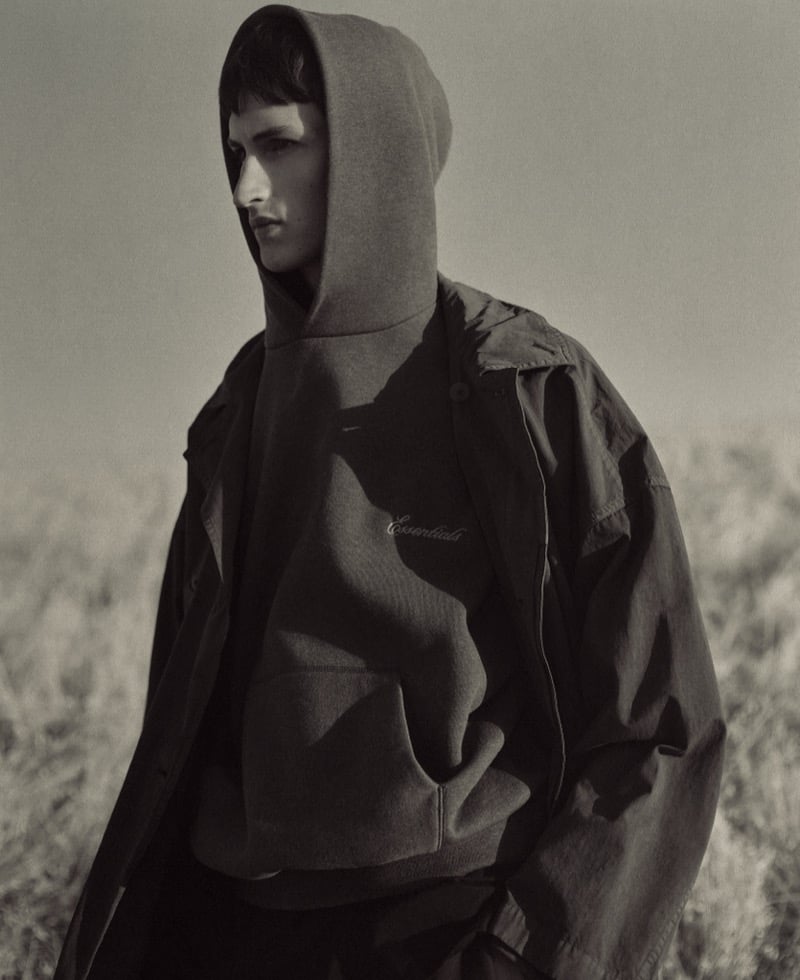 Anden Scudder in profile against a pale open field, the hood of an ESSENTIALS zip sweatshirt drawn up over his head beneath an oversized dark jacket and a longer coat, the Essentials script visible on the chest