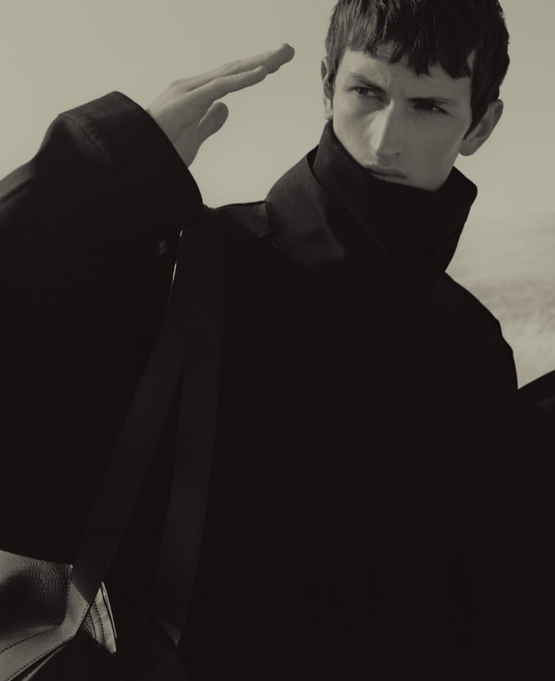 Anden Scudder raising one hand to his forehead, wearing a large dark funnel-neck coat with a leather tote bag strap over his shoulder, faint horizon behind him