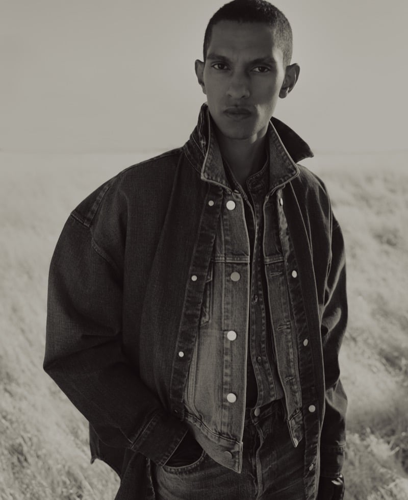Matthew Seymour with close-cropped hair standing in a dry field, wearing an oversized denim jacket layered over a denim trucker jacket and jeans, one hand in his pocket
