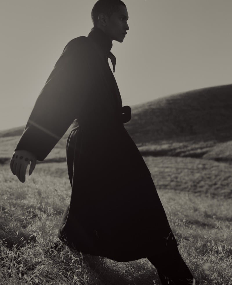 Matthew Seymour walking across a grassy hillside, backlit by low sun, wearing a long dark overcoat that falls past his knees, rolling hills in the background