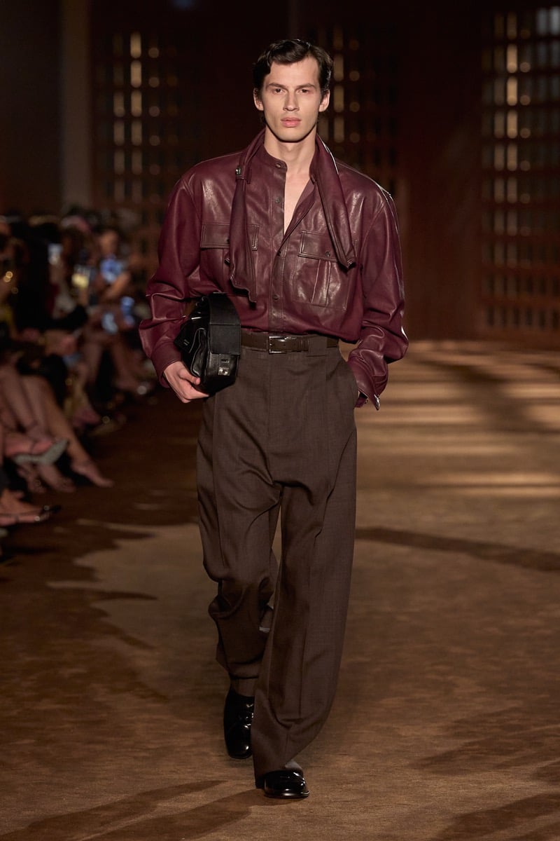 A model in a burgundy leather shirt with a scarf collar, pleated wide trousers, and a leather bag, for Ferragamo's spring-summer 2027 collection