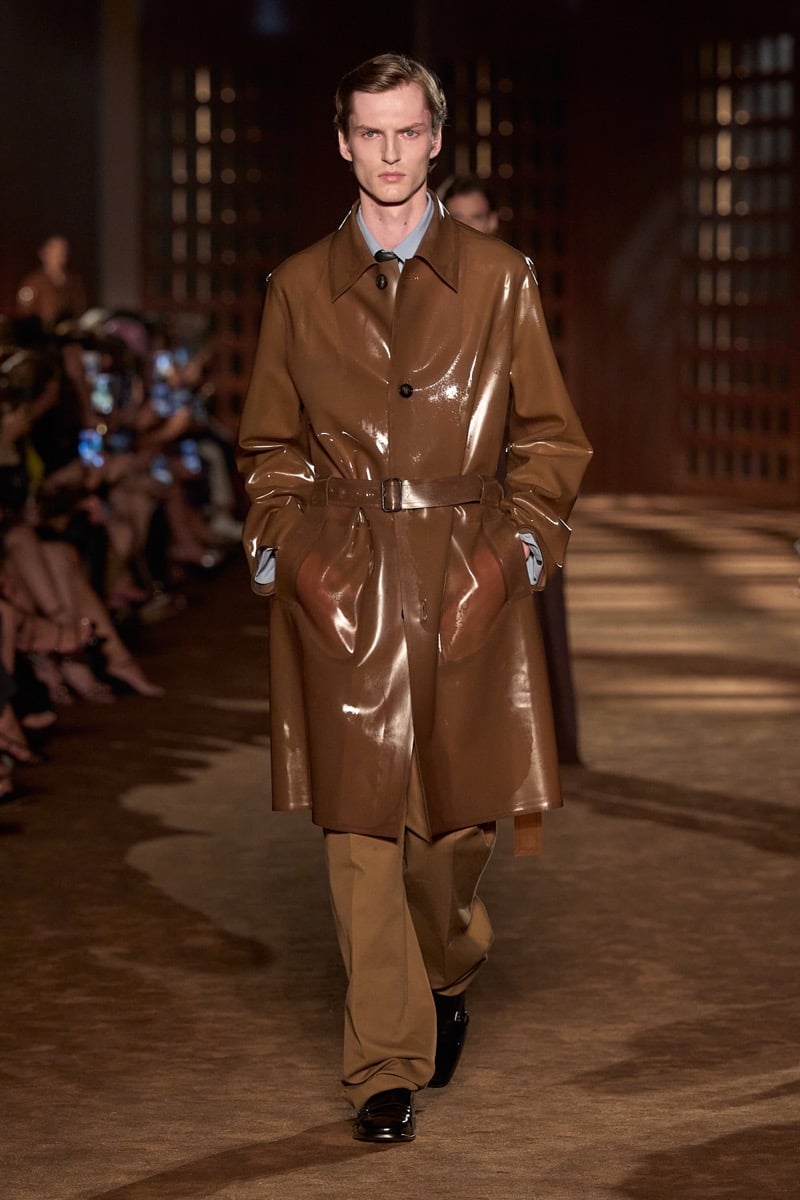 A model in a glossy brown rubber coat, belted at the waist, over a shirt and tie, for Ferragamo's spring-summer 2027 collection