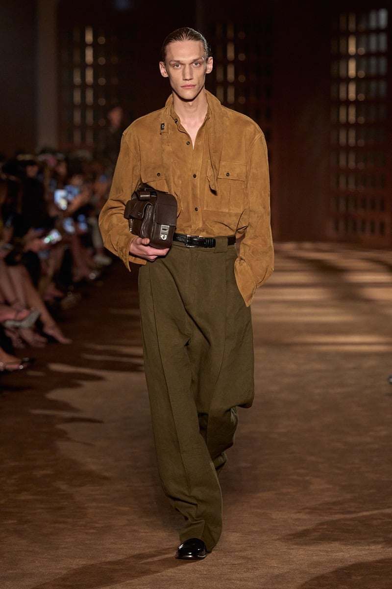 A model in a suede shirt jacket with a scarf collar and olive pleated trousers, holding a leather bag, for Ferragamo's spring-summer 2027 collection