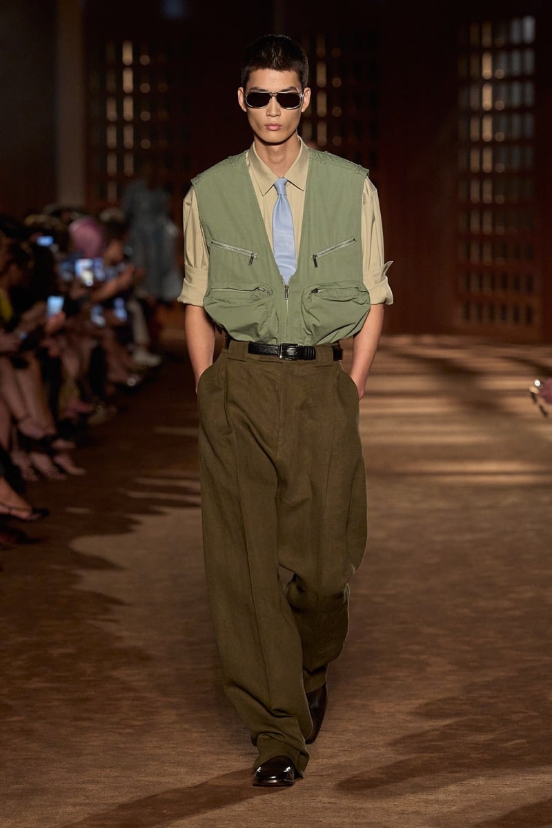 A model in aviator sunglasses and a green utility vest over a shirt and tie, with olive pleated trousers, for Ferragamo's spring-summer 2027 collection