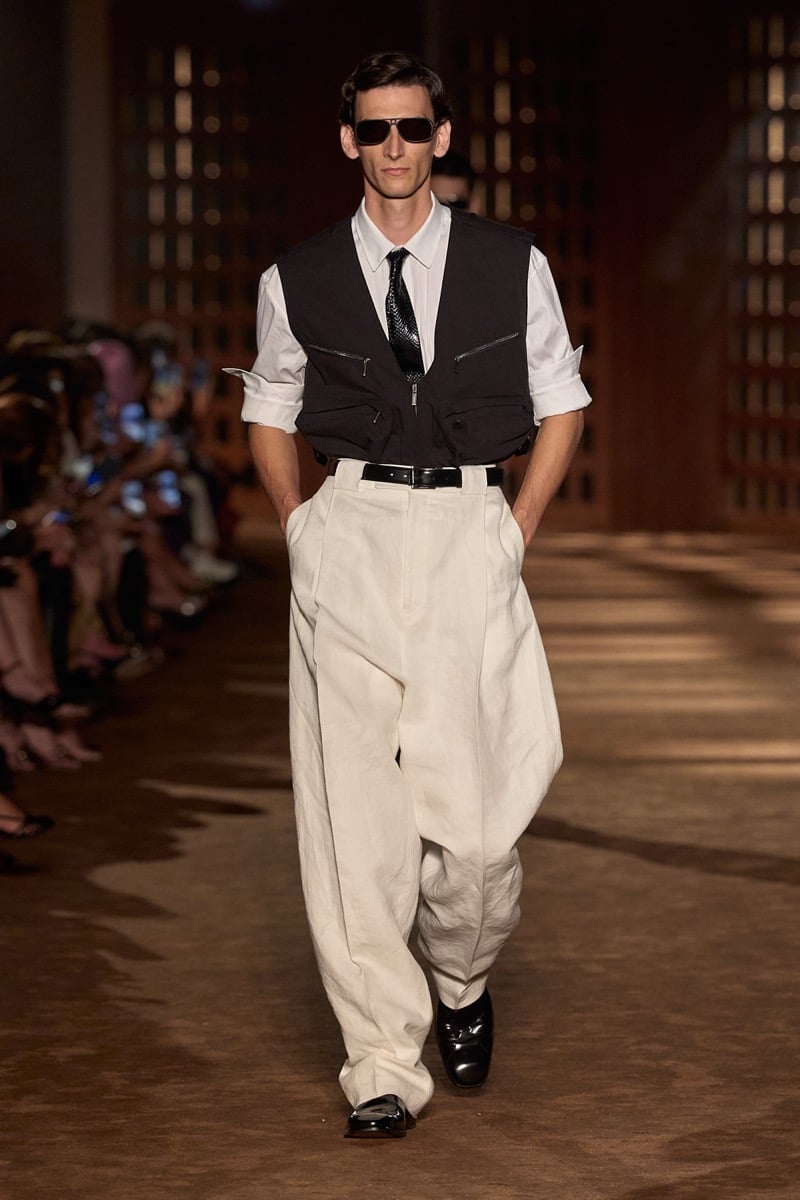 A model in aviator sunglasses and a zip-pocket vest over a shirt and tie, with pleated linen trousers, for Ferragamo's spring-summer 2027 collection