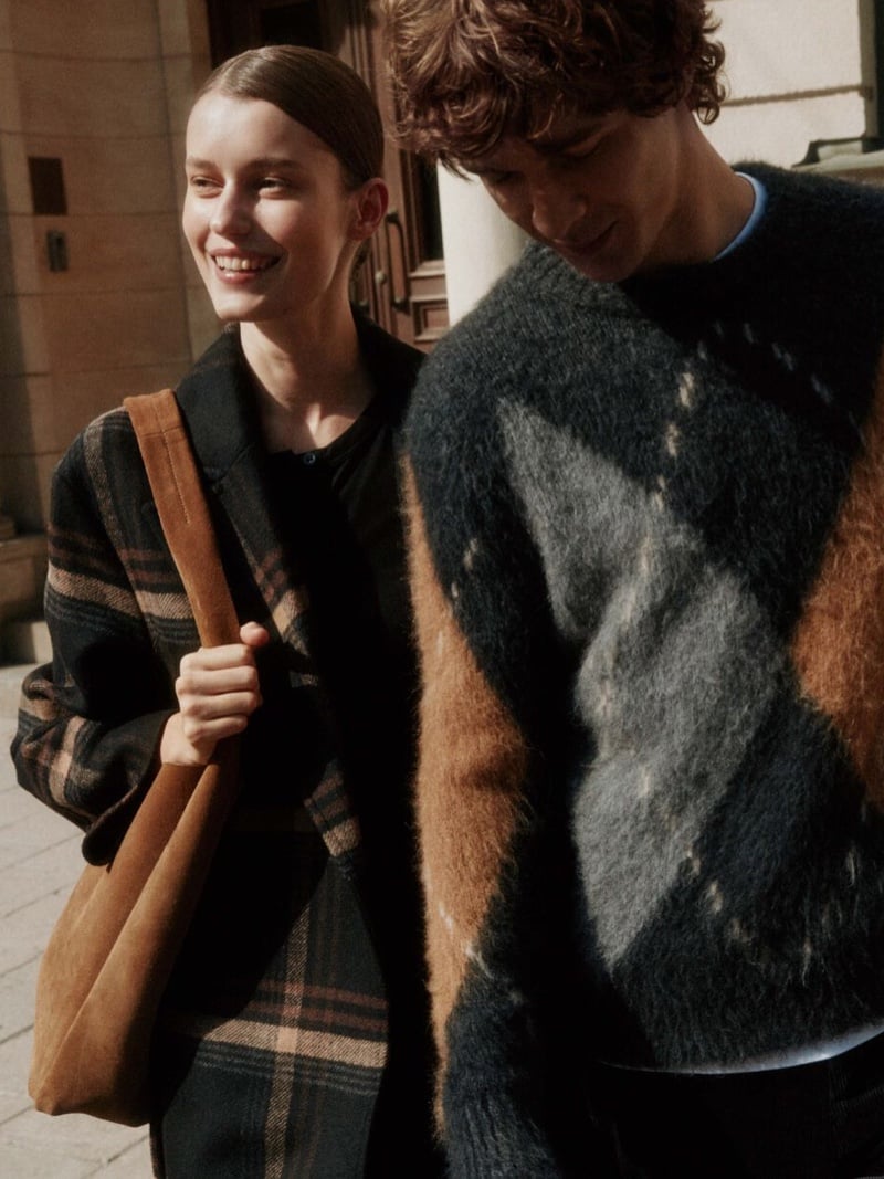 Efraim Schröder walks in sunlight wearing a brushed alpaca sweater with a large diamond intarsia pattern, next to Lesia Havrylets in a checked jacket with a suede tote bag on her shoulder
