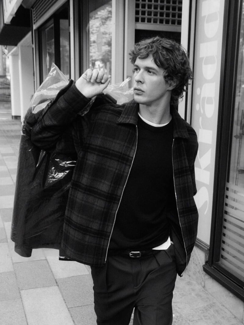 Black-and-white photo of Efraim Schröder walking a city sidewalk with dry cleaning in plastic slung over one shoulder, wearing a checked zip-front blouson jacket over a dark crew-neck knit and trousers