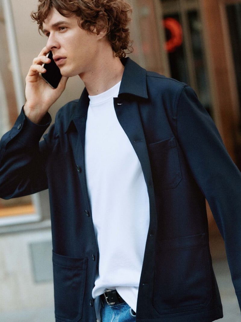 Efraim Schröder holds a phone to his ear while walking, wearing an unbuttoned navy overshirt with patch pockets over a white T-shirt and blue jeans