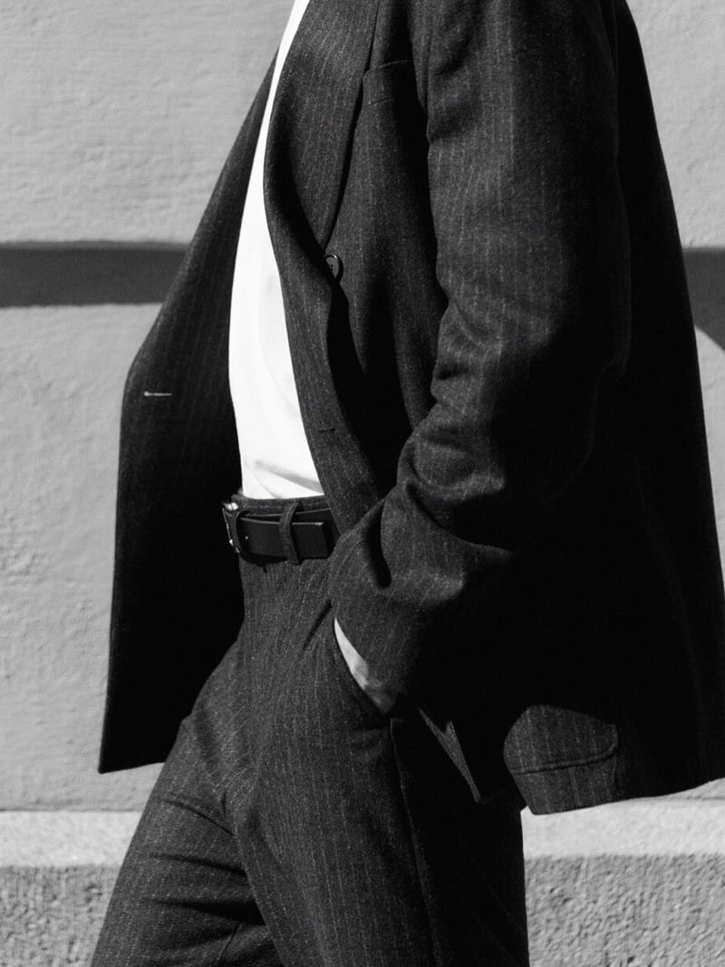 Black-and-white detail of Efraim Schröder mid-stride in a pinstripe double-breasted suit worn over a white T-shirt with a leather belt, cropped at the shoulders and knees