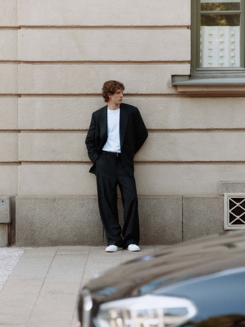 Efraim Schröder leans against a pale stone building wearing a pinstripe double-breasted suit with wide trousers over a white T-shirt and white low-top sneakers, both hands in his pockets