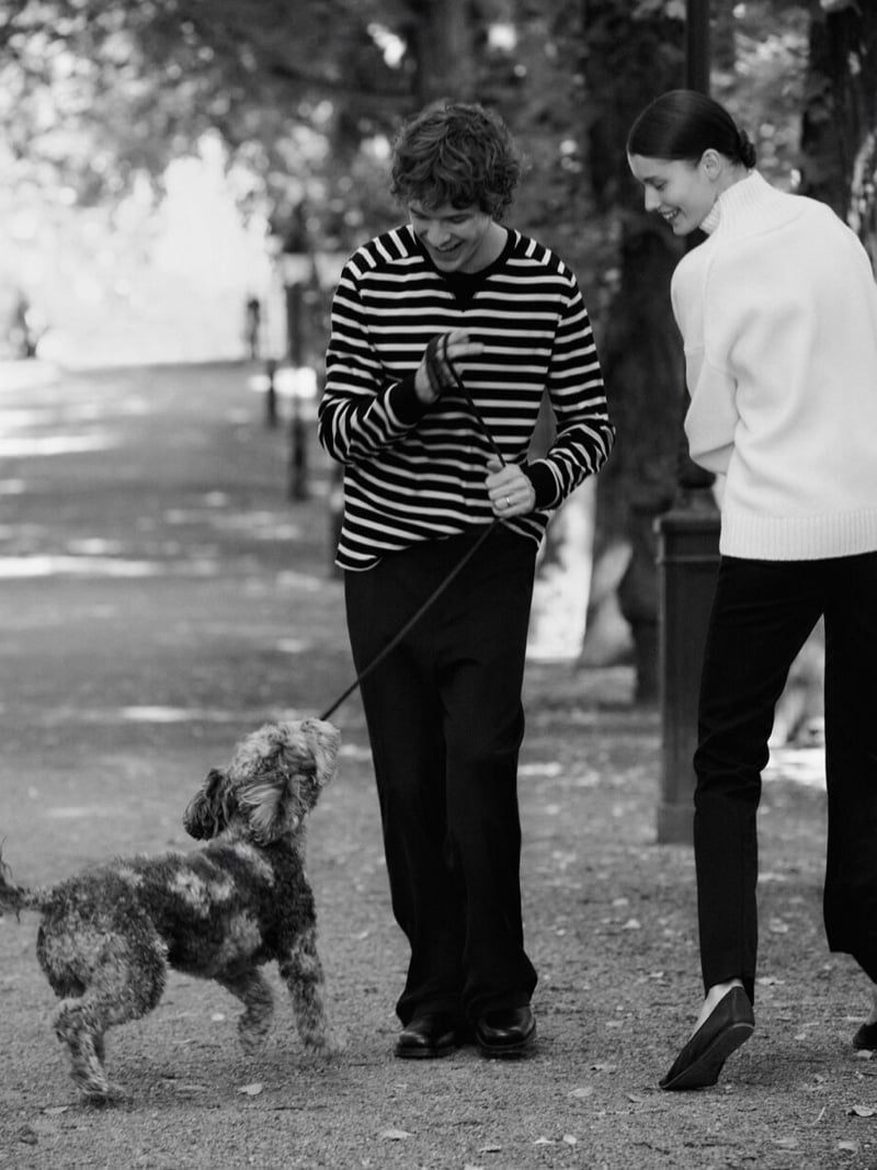 Black-and-white photo of Efraim Schröder in a horizontal-striped long-sleeve sweater and dark trousers walking a small curly-haired dog on a tree-lined path beside Lesia Havrylets, who wears a chunky roll-neck sweater