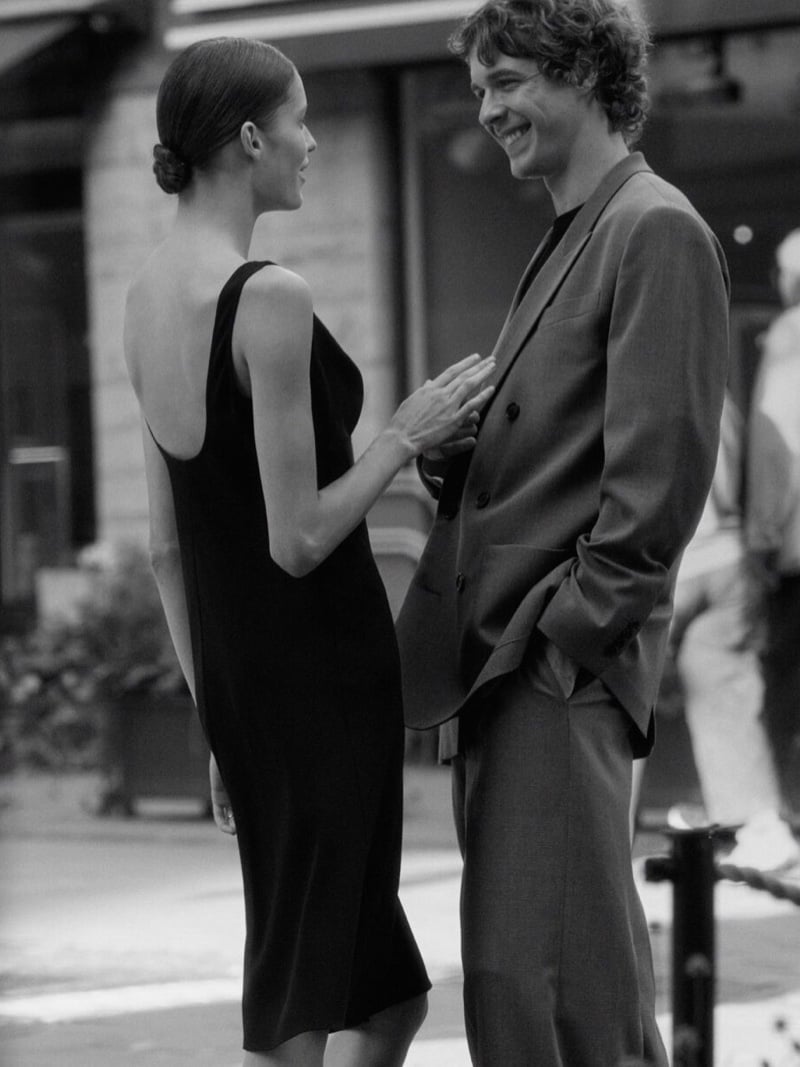 Black-and-white photo of Efraim Schröder and Lesia Havrylets facing each other on a busy sidewalk, he in a double-breasted suit and T-shirt, she in a scoop-back slip dress with a hand resting on his chest