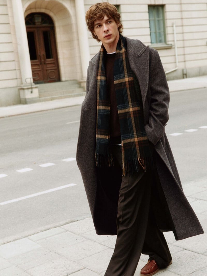 Efraim Schröder walks across a wide city street in an open double-breasted wool coat over a brown sweater, with a tan and dark checked scarf, dark wide-leg trousers, and brown leather shoes, for Filippa K pre-fall 2026