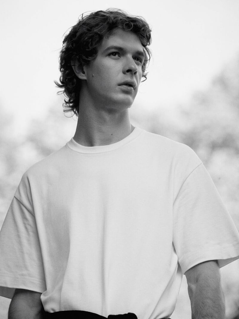 Black-and-white portrait of Efraim Schröder looking up and to the side, wearing an oversized crew-neck white T-shirt with blurred foliage behind him