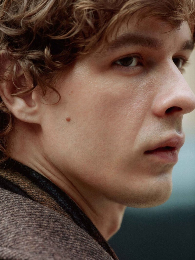 Extreme close-up of Efraim Schröder's face in profile, curly hair falling across his ear, the woven brown collar of a wool coat at his shoulder, for Filippa K pre-fall 2026