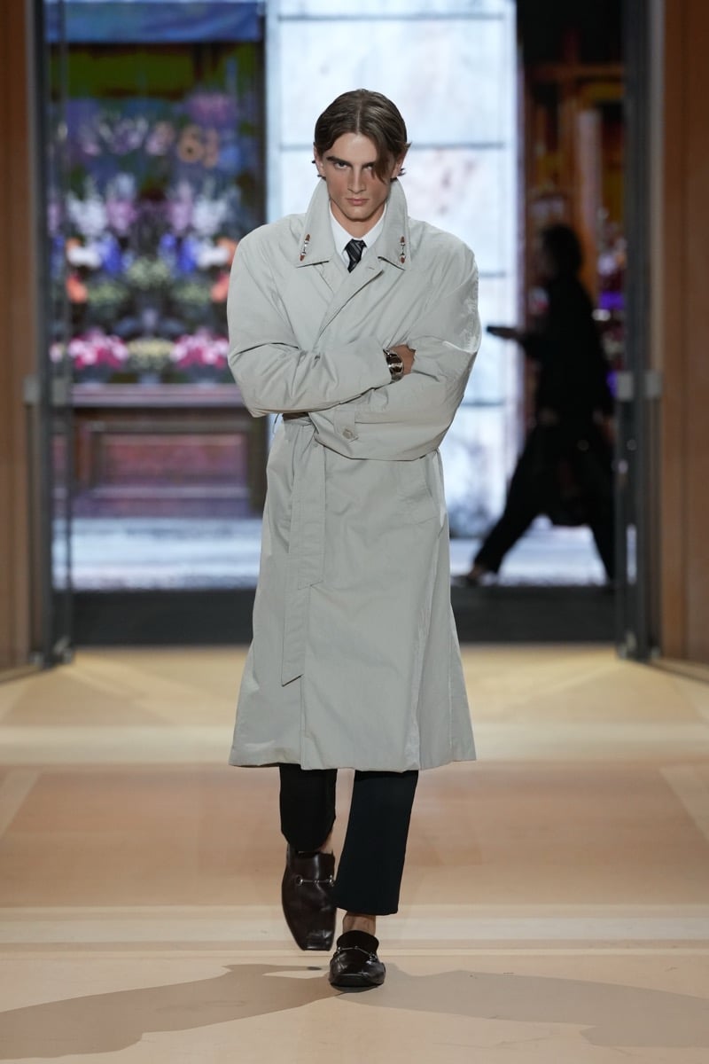 A model in a belted trench coat over a shirt and striped tie, with Horsebit loafers, for Gucci's spring-summer 2027 collection