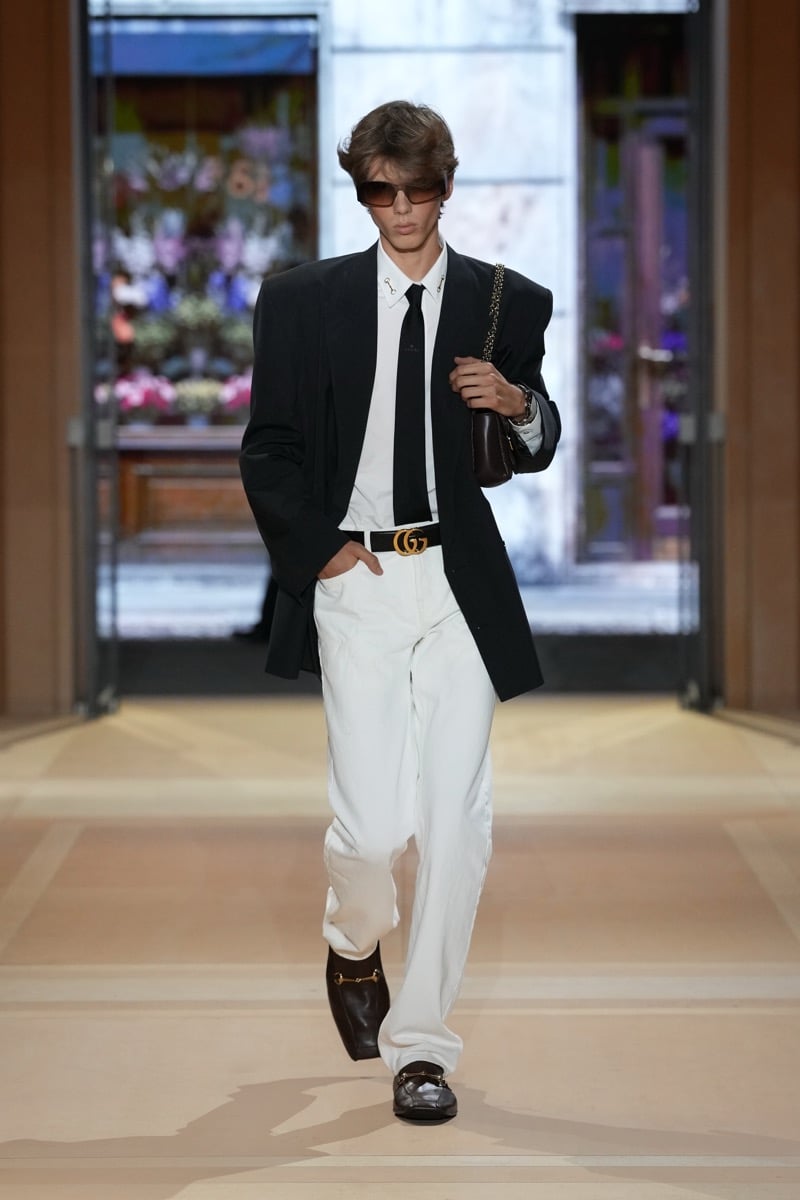 A model in a blazer over a shirt and skinny tie, with a GG belt, jeans, and a shoulder bag, for Gucci's spring-summer 2027 collection