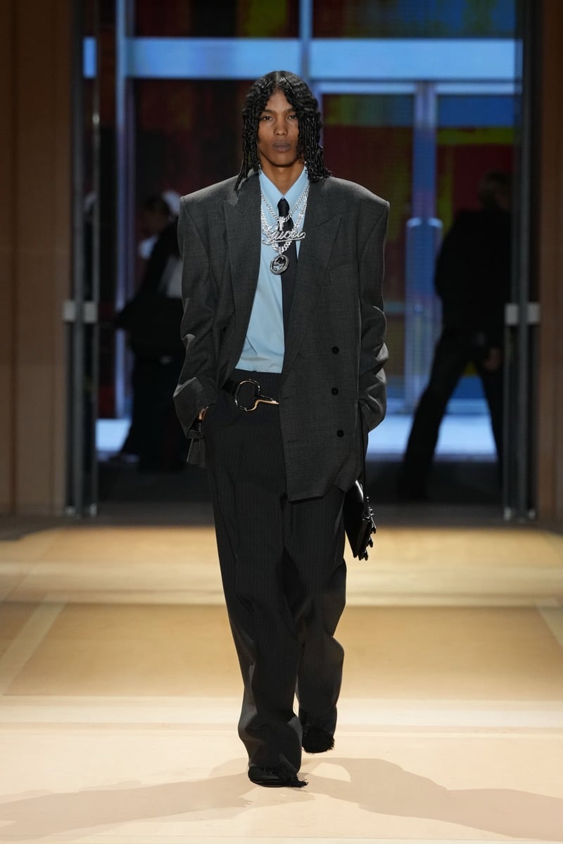 A model in a gray double-breasted blazer over a light blue shirt and tie, with a Gucci script chain and pinstripe trousers, for Gucci's spring-summer 2027 collection