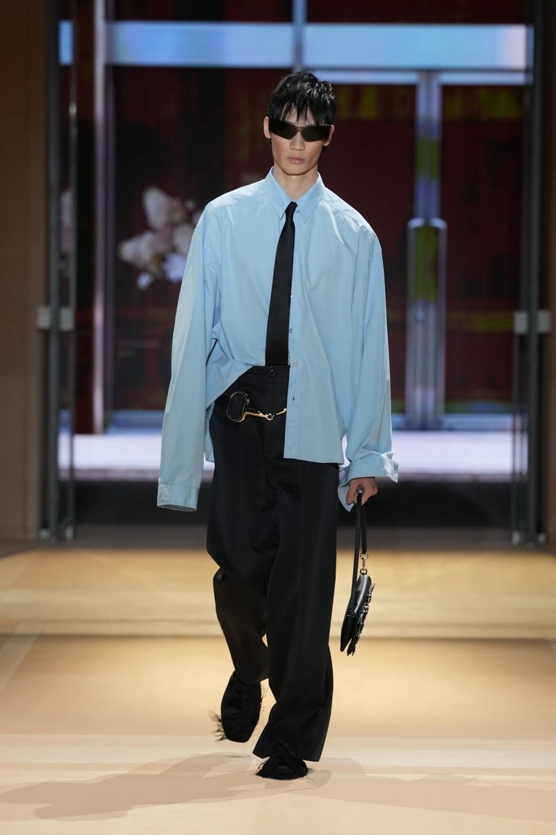 A model in an oversized light blue shirt and skinny tie, with a Horsebit belt and wide trousers, for Gucci's spring-summer 2027 collection