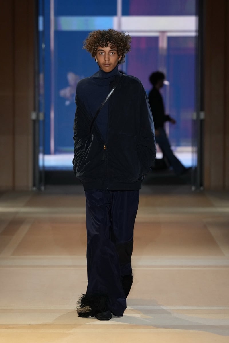 A model in a zip bomber over a turtleneck, with a crossbody strap, wide trousers, and shaggy slippers, for Gucci's spring-summer 2027 collection