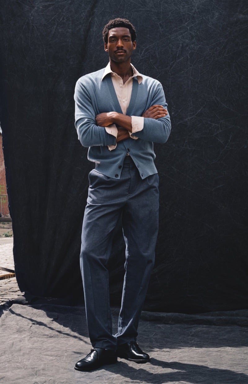 Halid Nuhu stands with arms crossed in a blue cashmere cardigan over a collared shirt with pleated trousers and black leather shoes, for Todd Snyder's Essential Pieces