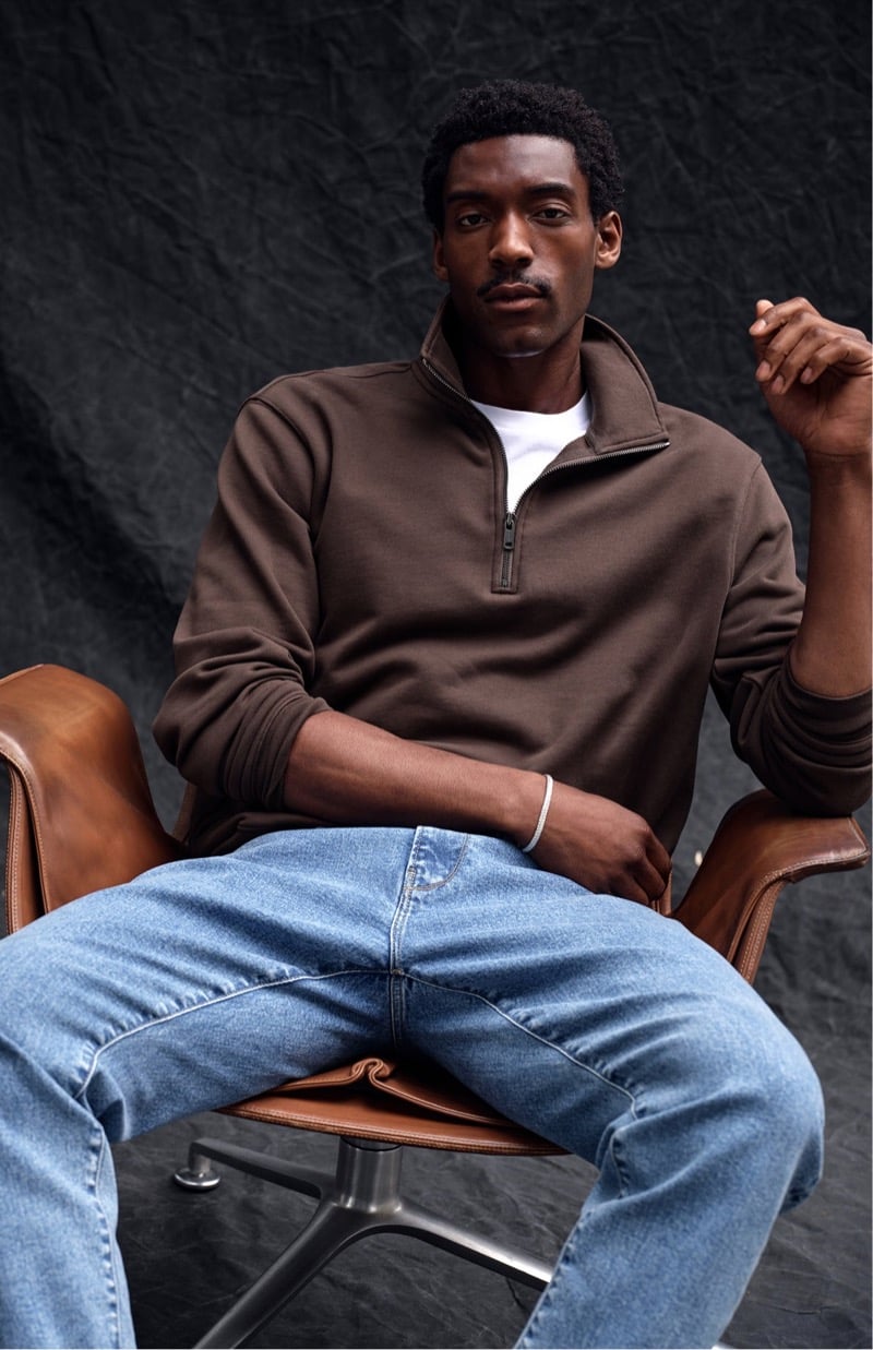Halid Nuhu sits in a leather chair in a brown quarter-zip sweatshirt over a tee with blue jeans, for Todd Snyder's Essential Pieces
