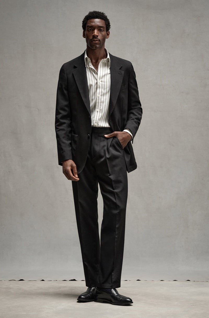 Halid Nuhu stands with a hand in his pocket in a dark Madison suit with an open striped shirt and black loafers, for Todd Snyder's Essential Pieces
