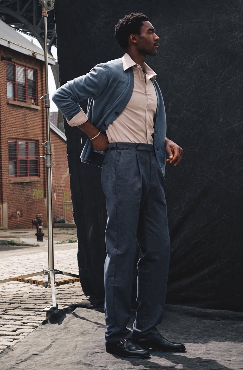 Halid Nuhu stands in profile with a hand on his hip in pleated side tab trousers, an open cardigan, and a collared shirt on a cobblestone set, for Todd Snyder's Essential Pieces